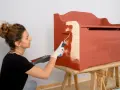 Mujer con guantes blancos pinta un mueble de madera con rodillo, cubriendo el barniz con pintura roja.