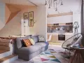 Un sofá gris con cojines coloridos. Detrás, una cama elevada y una pared con cuadros. La cocina moderna y una silla de diseño completan el espacio.