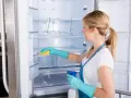 Mujer con guantes azules limpia el interior de una nevera vacía con una esponja amarilla y spray.