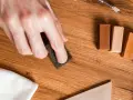 Mano lijando un parquet rayado con una esponja. Cerca, barras de cera para madera y un paño blanco.