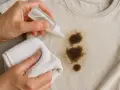 Manos aplicando líquido a una mancha de aceite en ropa beige, con un paño blanco listo para frotar. Aprende como quitar manchas de aceite de la ropa.
