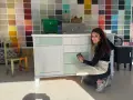 Mujer sonríe mientras lija un mueble para pintar. Detrás, una pared con muestras de colores.