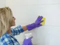 Mujer con guantes morados limpiando azulejos de cocina con esponja amarilla y spray.