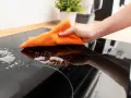 Mano limpiando vitrocerámica con paño naranja y espuma. Aprende como limpiar vitroceramica fácilmente.