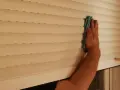Mano limpiando persianas por fuera con un paño azul.
