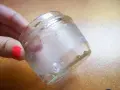 Mano con uña roja sostiene frasco de vidrio con residuos de pegamento. Aprende como quitar el pegamento de una pegatina.