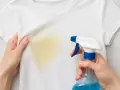Manos rocían una camiseta blanca con una mancha amarilla, mostrando cómo quitar manchas amarillas en seco.