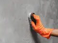Mano con guante naranja limpiando una pared de concreto con un cepillo, dejando círculos de espuma.