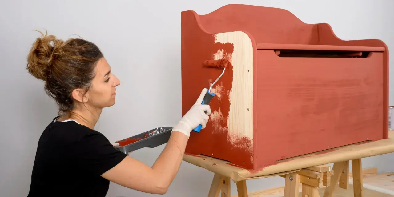 Mujer con guantes blancos pinta un mueble de madera con rodillo, cubriendo el barniz con pintura roja.