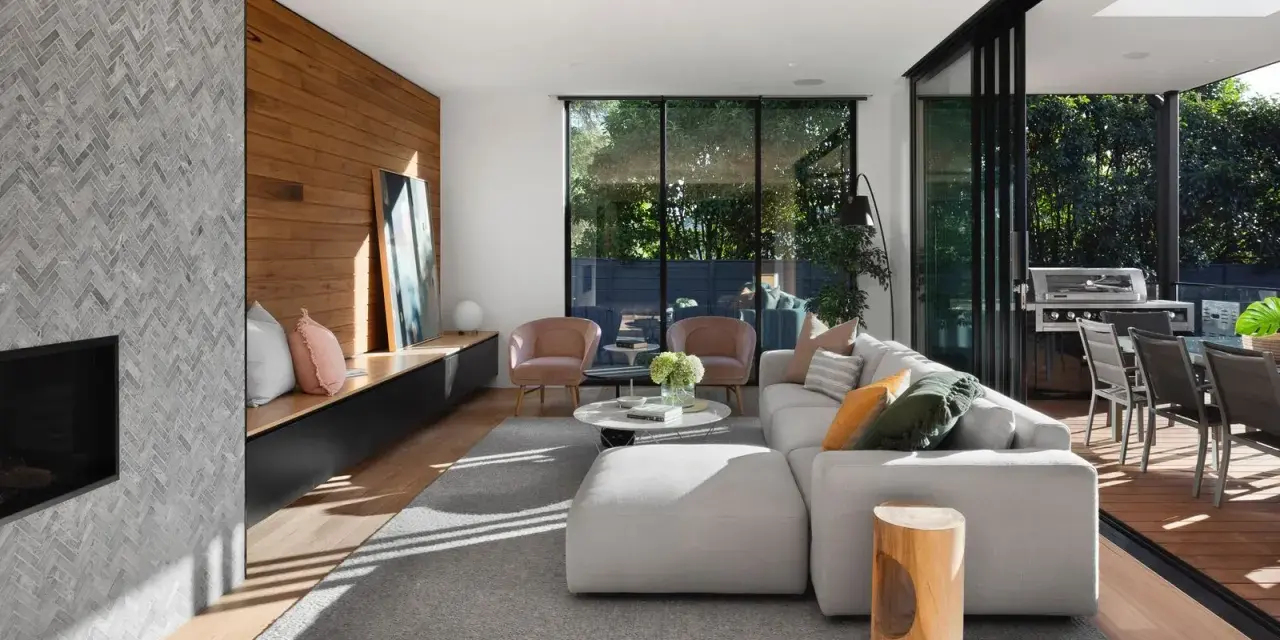Interior de casa de madera con chimenea de mosaico, sofá modular gris, sillones rosas y acceso a terraza con barbacoa.