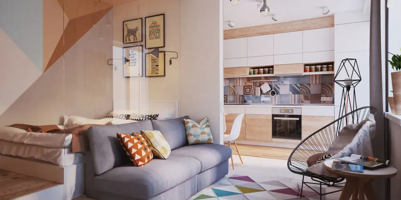 Un sofá gris con cojines coloridos. Detrás, una cama elevada y una pared con cuadros. La cocina moderna y una silla de diseño completan el espacio.