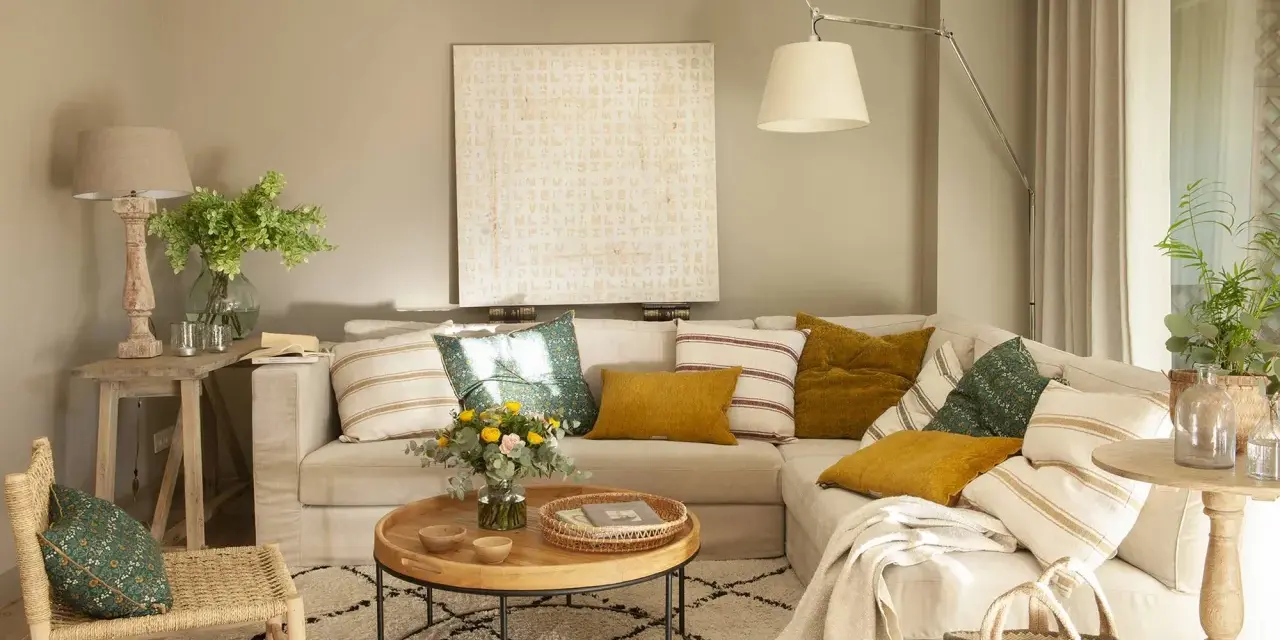Sofá beige con cojines variados, mesa de centro redonda de madera, alfombra estampada y una silla de mimbre. El color topo cual es, se ve en las paredes y cortinas.