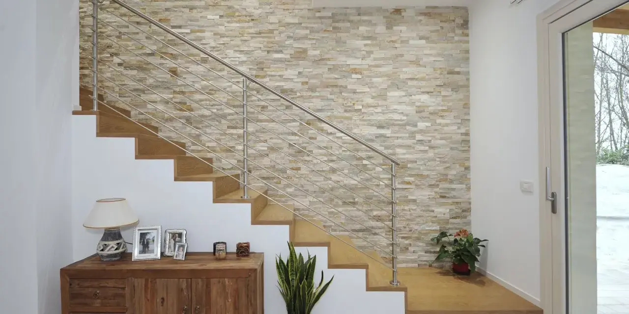 Elegante escalera con barandilla metálica y una pared de piedra natural para decorar pared subida escalera. Un mueble de madera y plantas completan la escena.