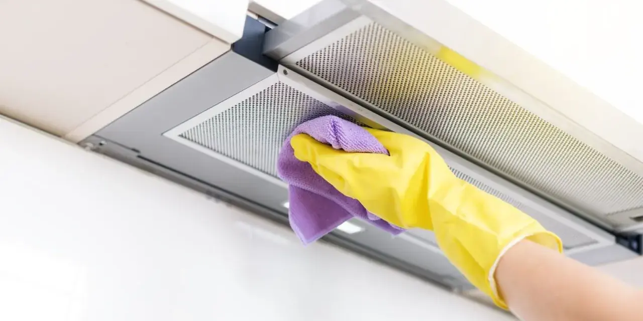 Mano con guante amarillo y paño morado limpiando el interior de la campana extractora, eliminando grasa y suciedad para un hogar reluciente.