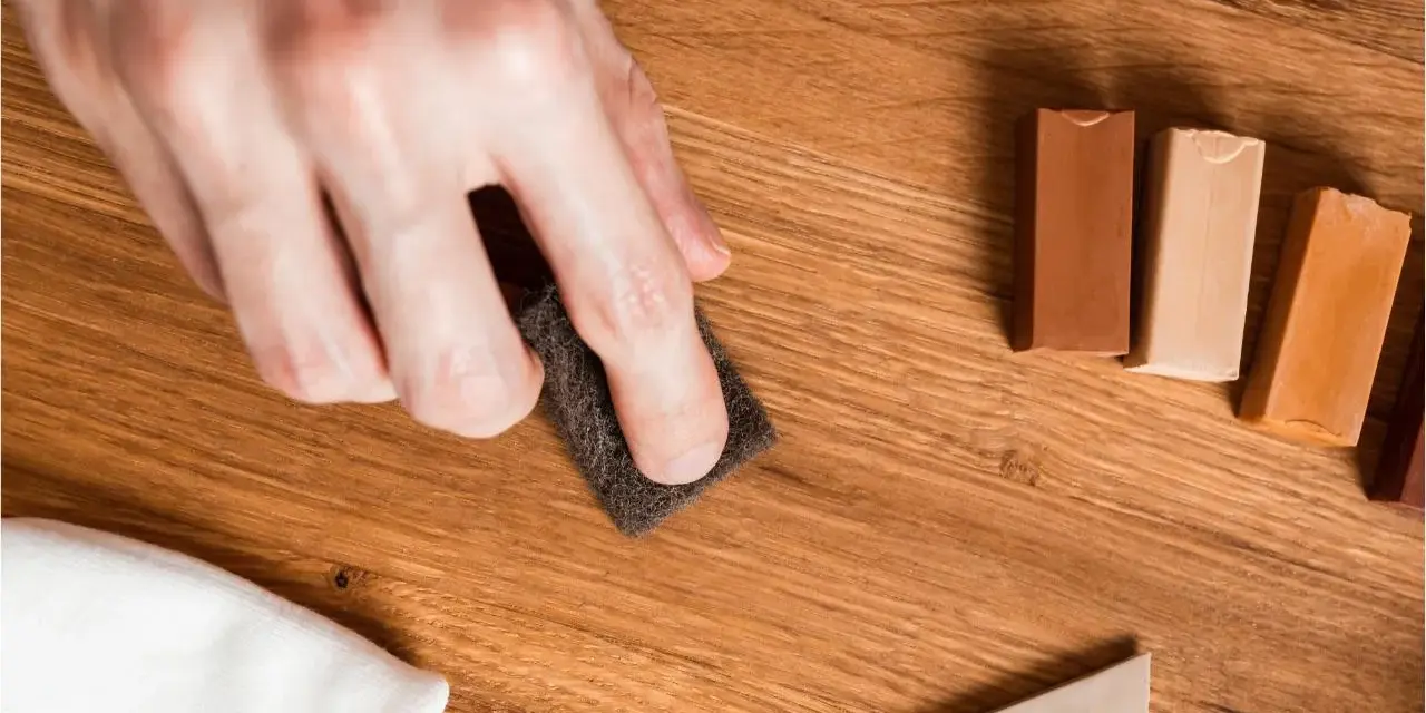 Mano lijando un parquet rayado con una esponja. Cerca, barras de cera para madera y un paño blanco.