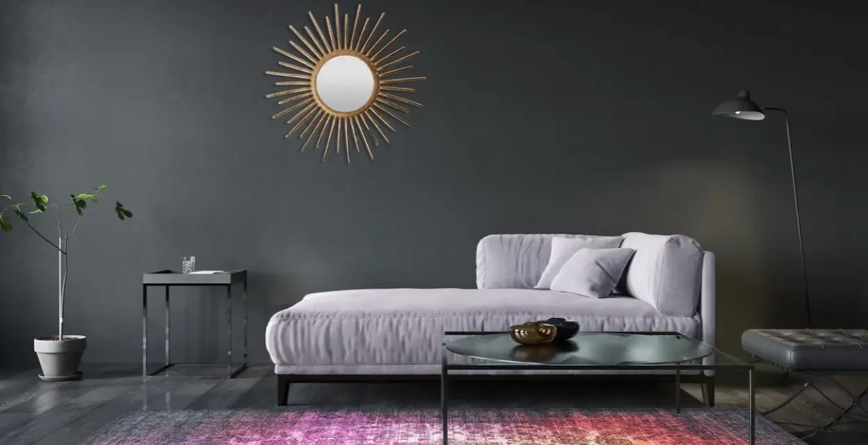 Sofá lila y alfombra vibrante sobre suelos oscuros. Un espejo sol dorado adorna la pared gris.