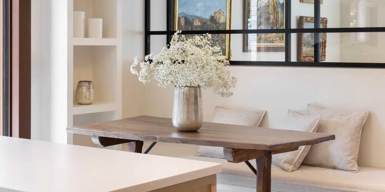 Un jarrón con flores blancas decora la mesa de madera rústica. El espacio alrededor de la mesa comedor es acogedor, con un banco tapizado y cojines.