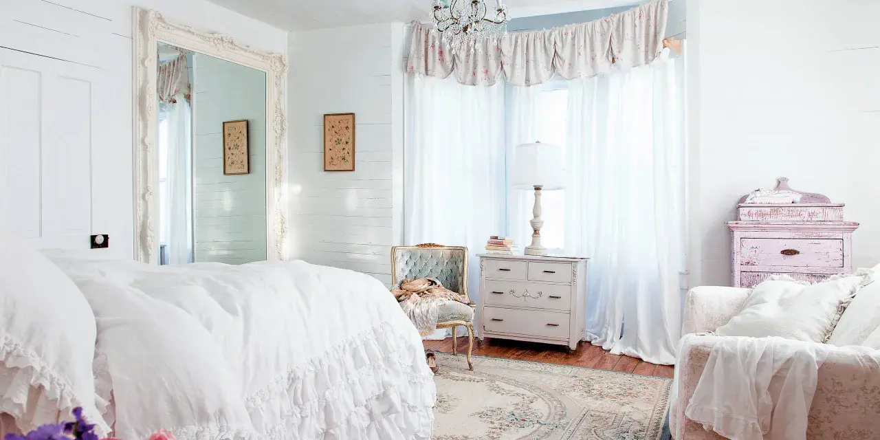 Dormitorio con cama blanca con volantes, espejo grande, cómoda antigua y un sillón. Estilo shabby chic.
