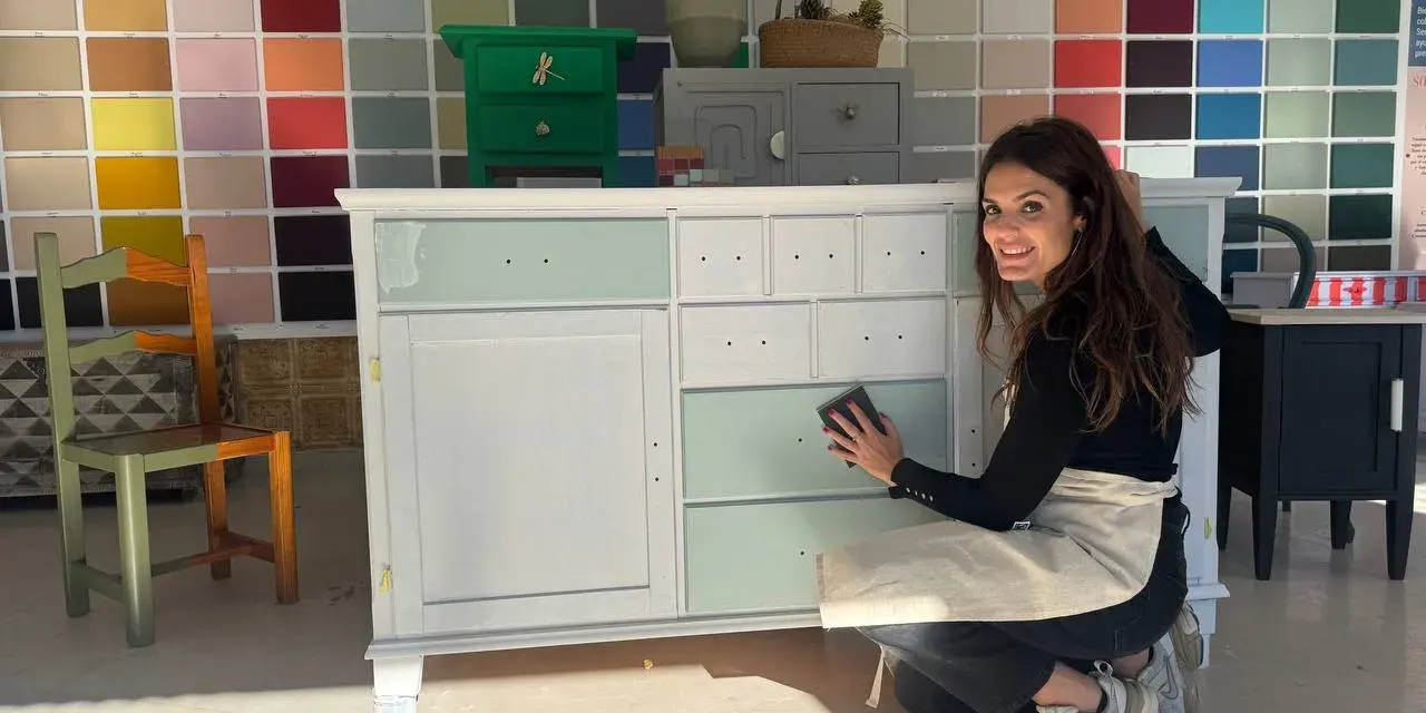 Mujer sonríe mientras lija un mueble para pintar. Detrás, una pared con muestras de colores.