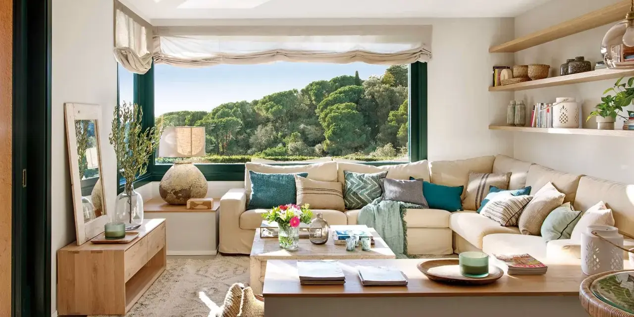 Acogedora decoracion salon comedor con sofá seccional beige, mesa de centro de madera y gran ventanal con vistas al bosque.