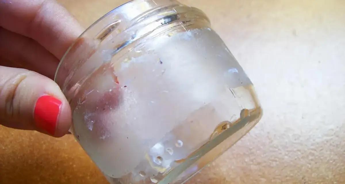 Mano con uña roja sostiene frasco de vidrio con residuos de pegamento. Aprende como quitar el pegamento de una pegatina.
