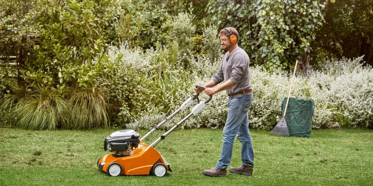Hombre con auriculares usa un escarificador naranja para airear el césped en un jardín verde.