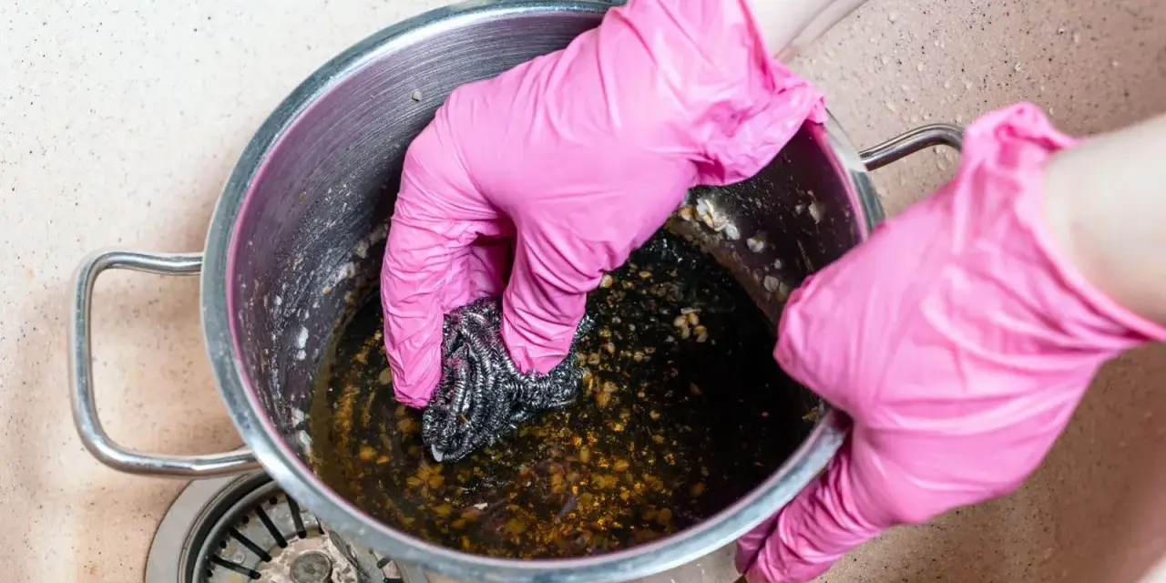 Manos con guantes rosas frotando una olla quemada con estropajo. Un reto para saber como quitar el quemado de una olla.