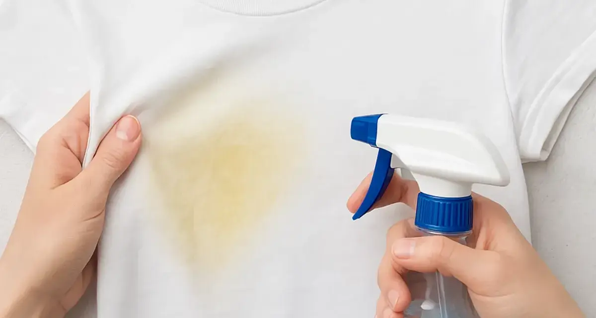 Manos rocían una camiseta blanca con una mancha amarilla, mostrando cómo quitar manchas amarillas en seco.