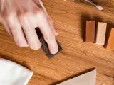 Mano lijando un parquet rayado con una esponja. Cerca, barras de cera para madera y un paño blanco.