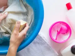 Manos lavando ropa con jabón en un recipiente azul. Aprende como quitar resina de la ropa con este método.