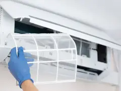 Mano con guante azul extrae filtro de aire acondicionado para limpiarlo por dentro.