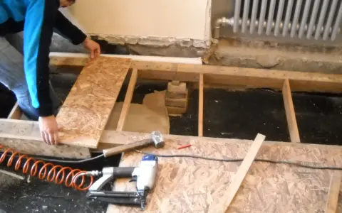 Hombre colocando tableros para nivelar suelo. Herramientas como martillo y clavadora neumática a la vista.