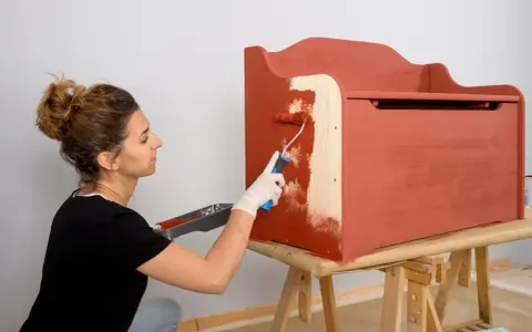 Mujer con guantes blancos pinta un mueble de madera con rodillo, cubriendo el barniz con pintura roja.