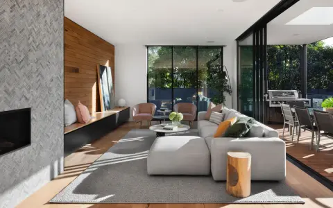 Interior de casa de madera con chimenea de mosaico, sofá modular gris, sillones rosas y acceso a terraza con barbacoa.