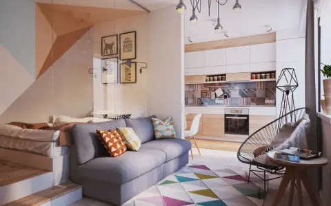 Un sofá gris con cojines coloridos. Detrás, una cama elevada y una pared con cuadros. La cocina moderna y una silla de diseño completan el espacio.