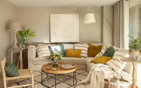 Sofá beige con cojines variados, mesa de centro redonda de madera, alfombra estampada y una silla de mimbre. El color topo cual es, se ve en las paredes y cortinas.