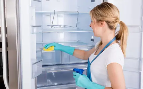 Mujer con guantes azules limpia el interior de una nevera vacía con una esponja amarilla y spray.