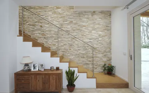 Elegante escalera con barandilla metálica y una pared de piedra natural para decorar pared subida escalera. Un mueble de madera y plantas completan la escena.