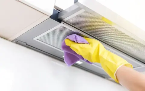 Mano con guante amarillo y paño morado limpiando el interior de la campana extractora, eliminando grasa y suciedad para un hogar reluciente.