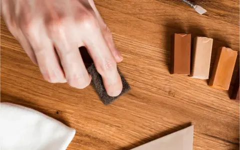 Mano lijando un parquet rayado con una esponja. Cerca, barras de cera para madera y un paño blanco.