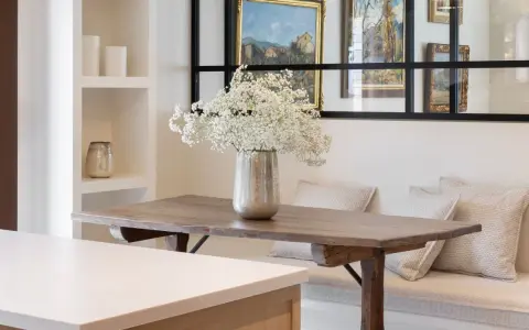 Un jarrón con flores blancas decora la mesa de madera rústica. El espacio alrededor de la mesa comedor es acogedor, con un banco tapizado y cojines.