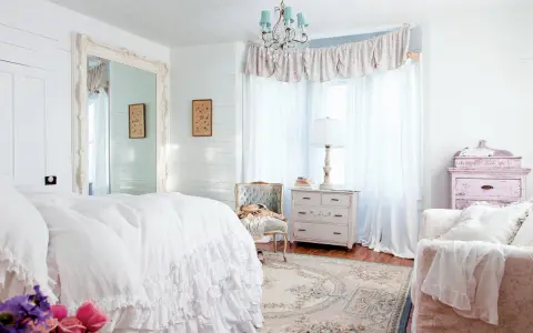 Dormitorio con cama blanca con volantes, espejo grande, cómoda antigua y un sillón. Estilo shabby chic.