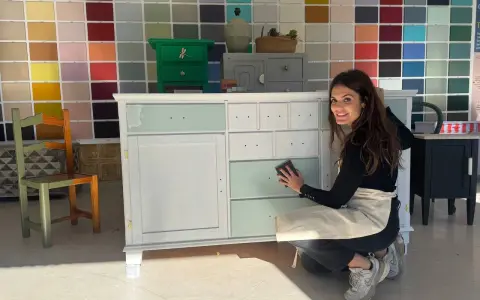 Mujer sonríe mientras lija un mueble para pintar. Detrás, una pared con muestras de colores.