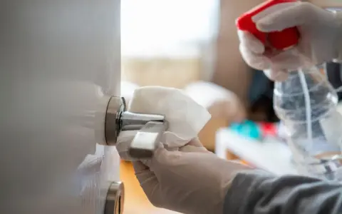 Manos enguantadas limpian un picaporte de puerta lacada en blanco con un paño.