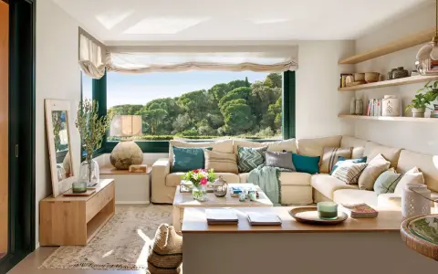 Acogedora decoracion salon comedor con sofá seccional beige, mesa de centro de madera y gran ventanal con vistas al bosque.