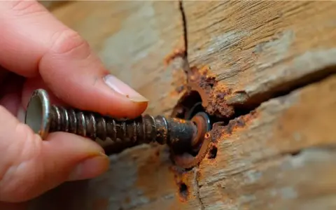 Mano girando un tornillo oxidado en madera agrietada. Se desatornilla hacia la izquierda.