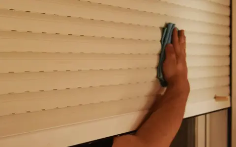 Mano limpiando persianas por fuera con un paño azul.