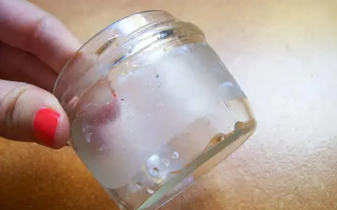 Mano con uña roja sostiene frasco de vidrio con residuos de pegamento. Aprende como quitar el pegamento de una pegatina.