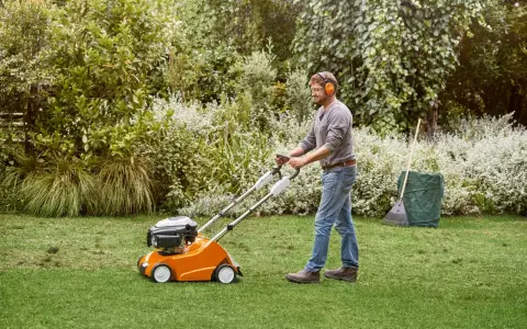 Hombre con auriculares usa un escarificador naranja para airear el césped en un jardín verde.