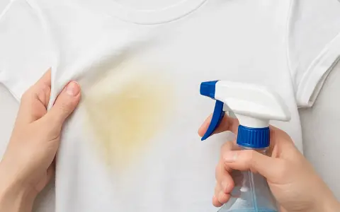 Manos rocían una camiseta blanca con una mancha amarilla, mostrando cómo quitar manchas amarillas en seco.